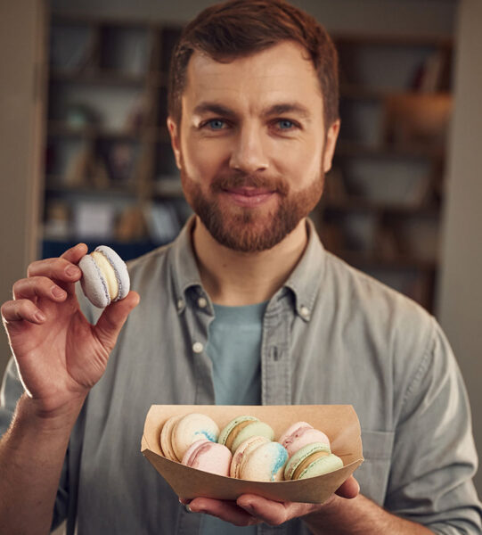 Holding and showing macaron. Man in casual clothes is holding sweets that are in the paper eco box.