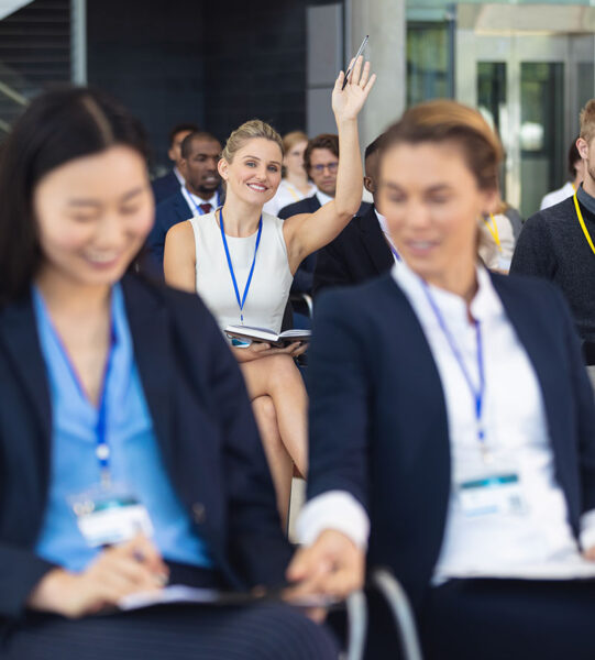 Front view of Young Caucasian businesswoman asking question in speech in conference room. With executives in the background.